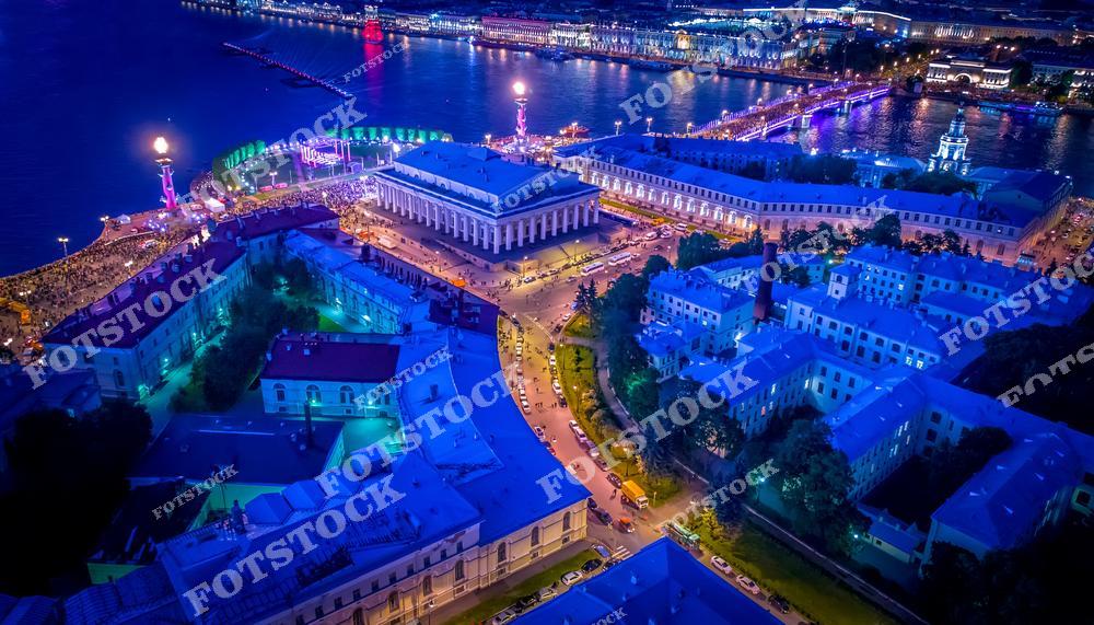 На фотографии изображена Стрелка Васильевского острова в ночное время, во время празднования, когда площадь заполнена людьми. Ростральные колонны и здание Биржи освещены яркой иллюминацией, придавая торжественный вид всему ансамблю. Ночной Санкт-Петербург сияет разноцветными огнями, которые отражаются в водах Невы.